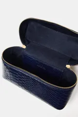 Bolso tipo vanity de color azul marino con textura de piel de serpiente en relieve. Cuenta con un asa de mano rígida metálica en tono dorado, cierre de doble cremallera y correa bandolera extraíble y ajustable.