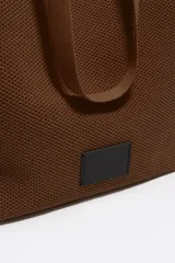 Bolso shopper de estructura blanda y gran tamaño, confeccionado en tejido técnico tipo mesh. Cuenta con doble asa de hombro, cierre mediante cordón ajustable y etiqueta de cuero con logo grabado en la parte frontal.