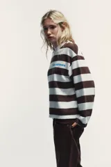 Camiseta de manga larga con cuello redondo y estampado de rayas horizontales en blanco y marrón. Incluye texto azul "TRIBECA MIDTOWN" estampado en el pecho.