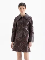 Chaqueta de cuero vacuno color marrón con acabado brillante, diseño de cuadros con líneas finas en tonos rojizos y beige, y detalle de volados de cuero aplicados en forma de trama. Posee cuello camisero, cierre frontal con botones y mangas largas.