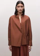 Camisa de algodón color marrón, de diseño cruzado estilo sobre con lazo lateral para anudar, cuello camisero, manga larga y calce amplio.