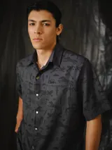Camisa de manga corta color gris oscuro con estampa exclusiva de símbolos, animales y elementos de la marca en tono negro. Presenta cuello camisero, dos bolsillos delanteros con solapa, corte relajado y cierre frontal con botones metálicos a presión.