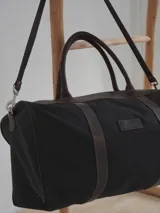 Bolso de viaje tipo duffel de lona negra con detalles en cuero tostado, asas de mano y correa ajustable para el hombro.