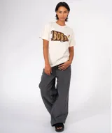 Remera color crudo con cuello redondo y manga corta. Estampado frontal de una banderín marrón con la palabra "BOSTON" en letras doradas con efecto glitter. Incluye dos pines decorativos prendidos en la zona superior derecha del pecho.