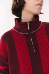 Buzo de tejido de punto con diseño de rayas verticales en tonos bordó y rojo. Presenta cuello alto con cierre frontal, mangas largas con coderas en contraste y detalles de costuras visibles.
