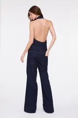 Enterito largo confeccionado en denim azul, sin mangas, con cuello halter y solapas de camisa, cierre frontal con botones metálicos y bolsillos frontales con solapa. Presenta pantalón de corte amplio.