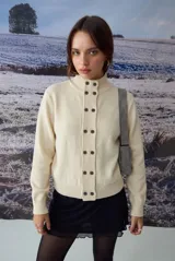 Cardigan de punto color crema con cuello alto y cierre frontal de botones oscuros. Presenta un diseño de textura acanalada en la franja central de los botones y mangas largas.
