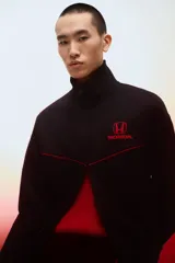 Campera deportiva negra de efecto neopreno con cierre bidireccional, cuello elevado y bolsillos laterales. Presenta detalles de ribetes rojos en el pecho y el logo de Honda bordado en rojo.