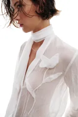 Blusa de tejido semitransparente con efecto arrugado, escote en pico y manga larga. Presenta un detalle decorativo frontal con estructura en forma de flor.