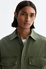 Camisa cargo color verde militar de corte regular, confeccionada en tejido ripstop de algodón. Cuenta con cuello inglés, cierre frontal de botones, bolsillos con solapa en el pecho y mangas largas con puños abotonados.