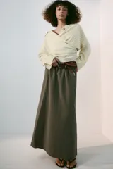 Falda midi tipo capa de tiro medio, confeccionada en mezcla de algodón color verde militar. Cuenta con cintura con trabillas, bolsillos de vivo en los laterales, cierre lateral con cremallera oculta y aplicación de cinturón marrón de estilo western con hebilla metálica grabada.