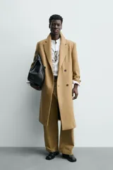 Abrigo largo color camel, de corte relajado (relaxed fit), confeccionado en tejido con mezcla de lana. Presenta cuello con solapas de muesca, manga larga, bolsillos de vivo en cadera, bolsillo interior y cierre frontal de botonadura cruzada.