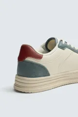Zapatilla deportiva de estilo retro con diseño de paneles en combinación de colores crema, verde azulado y detalle en rojo en el talón. Presenta suela con volumen y cierre con cordones.
