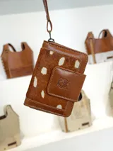Mini billetera de cuero con diseño de pelo animal print en tonos marrón y blanco. Presenta un cierre de solapa con broche y un logo de búho grabado en el frente, además de una correa de muñeca extraíble.