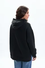 Canguro deportivo de color negro, con capucha ajustable mediante cordones, mangas largas y corte holgado.