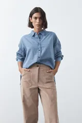 Camisa de jean en tencel con silueta amplia, hombros caídos, cuello clásico y cierre frontal con botones. Presenta ruedo curvo con tajos laterales y pliegue en la espalda.