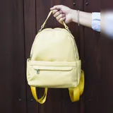 Mochila de tamaño mediano en color amarillo, confeccionada en símil cuero. Presenta un compartimento principal con cierre metálico, un bolsillo frontal con cierre y broche, y bolsillos laterales. Incluye correas regulables para la espalda, asa de mano y una correa adicional para llevar al hombro.