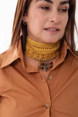 Choker tejido a mano en crochet color mostaza, con diseño de red y borde festoneado. Presenta una cadena dorada en la parte inferior de la cual cuelgan múltiples dijes de cuentas esféricas en tonos tierra y verde.