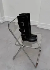 Botas de caña alta confeccionadas en ecocuero negro, con diseño de tres hebillas metálicas laterales y suela plana.