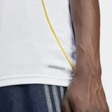 Camiseta de fútbol Adidas del Real Madrid, modelo Primera Equipación temporada 25/26, para hombre. Diseño blanco con detalles en negro (logo Adidas, tres franjas en hombros, texto del sponsor) y ribetes laterales en color naranja/dorado. Incluye escudo del club bordado y logo de sponsor en manga.