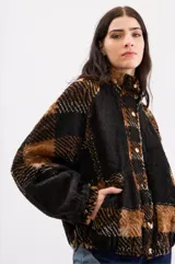 Campera tipo bomber oversize de paño italiano, con diseño de cuadros en tonos negro y marrón. Presenta cuello alto, cierre frontal con botones metálicos, mangas tipo murciélago y bolsillos laterales.