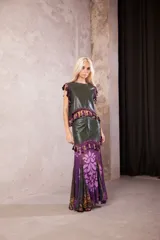 Falda larga de cuero verde oliva con diseño de paneles. La parte superior es de cuero liso con cierre frontal, decorada con un galón de borlas en tonos morados y verdes. La parte inferior presenta una falda de tela fluida con estampado floral y geométrico en tonos violeta, morado y verde.