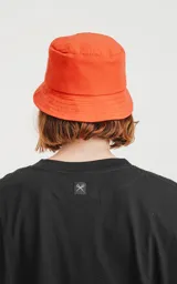 Gorro tipo bucket de tela color naranja vibrante, con un pequeño parche negro en el frente que tiene un logo bordado en forma de tijeras cruzadas.