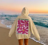 Canguro hoodie oversize color beige, con capucha, manga larga y gran diseño gráfico estampado en la espalda con flores rosas y texto en verde.