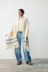 Chal tejido de punto en color crudo, con diseño artesanal que incluye un círculo amarillo y franjas horizontales en azul, con terminación de flecos en los extremos.
