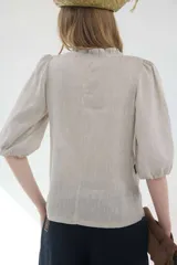 Camisa de lino color beige, de silueta romántica con escote a la base y mini volado. Cuenta con mangas 3/4 abullonadas con frunces y terminación en elástico. Posee detalles de puntillas negras en contraste en el frente y cierre frontal con botones ocultos bajo cartera.