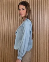 Camisa de denim de algodón ligero, con cuello en V adornado con grandes volados frontales y cierre de botones. Presenta mangas largas con puños elásticos fruncidos.