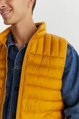 Chaleco puffer acolchado color mostaza, con cuello alto y cierre frontal. Diseño ligero y versátil con bolsillos laterales.