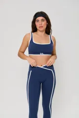 Conjunto deportivo compuesto por un top de escote cuadrado y calzas largas de tiro alto con diseño cruzado en la cintura. Ambas piezas son de color azul con detalles de ribetes blancos en los bordes y laterales. El top cuenta con el logo de la marca estampado en el frente.