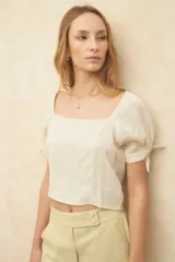 Top corto color blanco roto, con escote cuadrado, manga corta abullonada con lazos ajustables en el borde y textura arrugada en el tejido.