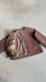 Chaqueta de gamuza color chocolate con detalles de bordado en color rojo, cuello redondo, cierre frontal con botones y mangas largas con puños abotonados.