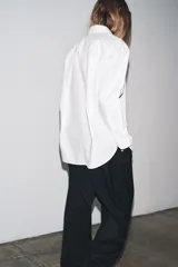 Camisa blanca de popelín de algodón 100%, con cuello solapa, manga larga terminada en puño y cierre frontal con botones ocultos por solapa. Presenta un corte holgado y ruedo curvo.