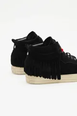 Zapatillas de caña media confeccionadas en cuero vacuno descarne color marrón. Presentan un diseño con flecos laterales, cordones al tono y un pequeño dije de corazón rojo en la parte superior de la lazada. Cuentan con suela de goma en color crema.