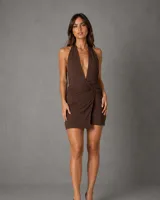 Vestido corto color chocolate con escote profundo tipo halter y espalda descubierta. Presenta un diseño con frunces drapeados en la parte frontal que realzan la silueta y una falda con efecto cruzado.