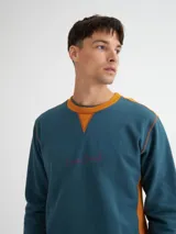 Buzo azul con cuello redondo, mangas largas y puños y bajo acanalados. Presenta bloque de color naranja en hombros y laterales, y estampado frontal con texto en color borgoña.