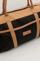 Bolso de viaje tipo duffel de lona negra con detalles en cuero tostado, asas de mano y correa ajustable para el hombro.
