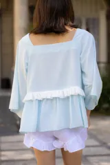 Blusa celeste de tela liviana con escote cuadrado, mangas tres cuartos amplias y detalle de volado horizontal en la parte frontal.