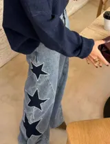 Jean de tiro alto con pierna amplia de corte recto, confeccionado en denim de lavado claro. Presenta aplicaciones de estrellas en contraste con bordes decorados con tachas metálicas.