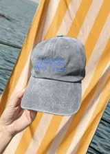 Gorro de visera de tela color gris desgastado, con bordado frontal en hilo rojo que dice "COSTA DULCE" y un diseño de ondas o símbolos repetidos encima.