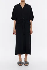 Vestido largo negro de viscosa con textura, corte recto, cuello camisero con solapas, manga corta, cierre frontal de botonadura, bolsillos plastrón en el pecho, bolsillos con tapeta en bajo delantero y cinturón de tejido.
