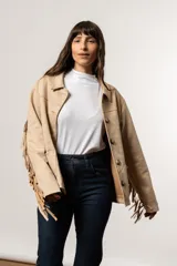 Chaqueta color beige de gamuza sintética con flecos en mangas y canesú, cuello camisero y cierre con botones.