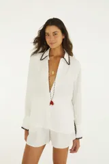 Camisa blanca de manga larga y calce relajado, confeccionada en tejido liviano de textura arrugada. Presenta cuello camisero, botonera frontal y detalles de ribete en zigzag marrón oscuro en cuello, puños y dobladillo.