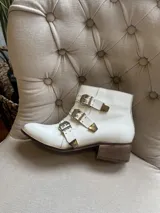 Bota corta blanca de cuero, con taco cuadrado de madera, punta fina y tres tiras cruzadas con hebillas metálicas plateadas y tachas decorativas.
