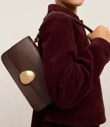 Cartera tipo shoulder bag de tamaño mediano, confeccionada en poliuretano color marrón oscuro. Presenta un diseño estructurado con tapa frontal y un distintivo detalle metálico dorado de forma orgánica en el cierre. Incluye asa larga ajustable para llevar al hombro.