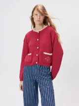 Cardigan de algodón grueso color rojo, con cuello redondo, cierre frontal de botones grandes y bolsillos cuadrados en el frente. Presenta detalles de pelo sintético en el cuello y los bolsillos, y logo bordado en uno de los bolsillos.