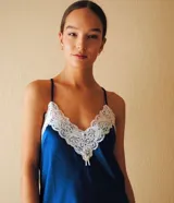 Camisón azul de satén elastizado con escote en V en la parte delantera y trasera, decorado con puntilla blanca. Tiene breteles finos y cruzados en la espalda, y un pequeño moño blanco en el centro del escote.